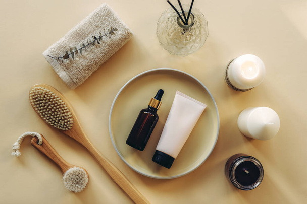 Cosmetic layout with serum bottle, cream tube, wooden brushes, candles and towel on beige background. Selfcare, home spa and relaxation concept. Flat lay, top view. - Photo, Image