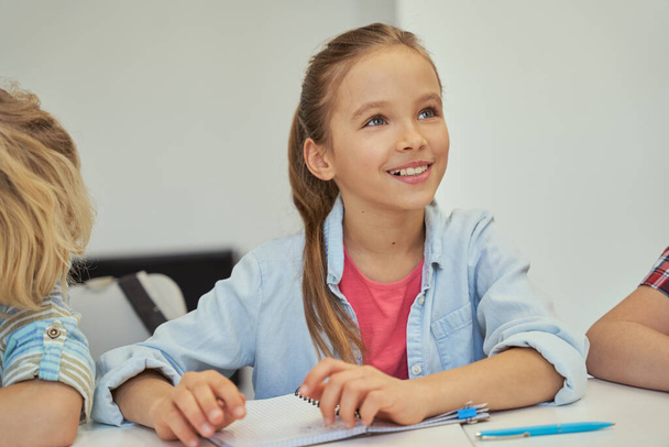 Retrato de una colegiala adorable sonriendo y escuchando mientras estudia, sentada a la mesa en el aula de la escuela primaria - Foto, imagen