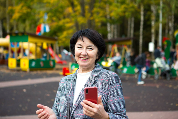 Portrait of a happy senora for 50 years in the park in autumn in a gray jacket on the playground. A woman laughs and uses a red smartphone - Valokuva, kuva