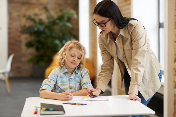 Supportive young female teacher in glasses helping little boy with the task. Kid studying in elementary school, sitting at the desk - Foto, Bild