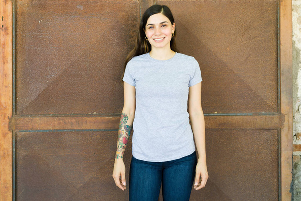 Усміхнена молода жінка носить сіру футболку на відкритому повітрі і показує нетиповий принт спереду
 - Фото, зображення