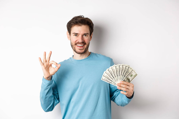 Schöner junger Mann zeigt schnelle Kredite Geld, machen okay Geste und lächelt mit Bargeld, steht über weißem Hintergrund - Foto, Bild
