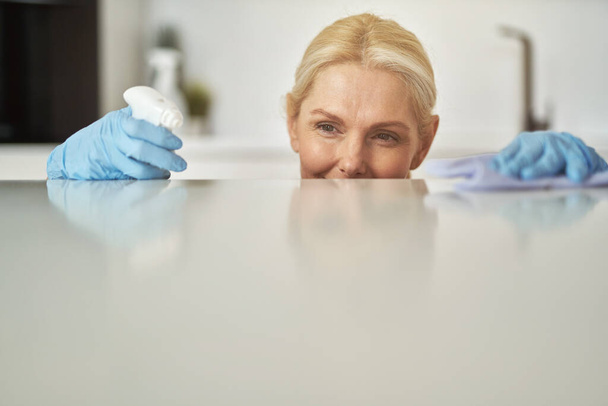 Porträt der attraktiven blonden Hausfrau in Gummihandschuhen, die die Sauberkeit der Oberfläche überprüft, während sie in der Küche mit Tuch und Spülmittel aufräumt - Foto, Bild