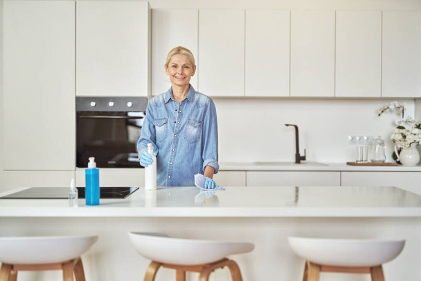 Uśmiechnięta dojrzała gospodyni domowa w gumowych rękawiczkach patrząca na kamerę podczas sprzątania powierzchni kuchni detergentem w domu w ciągu dnia - Zdjęcie, obraz