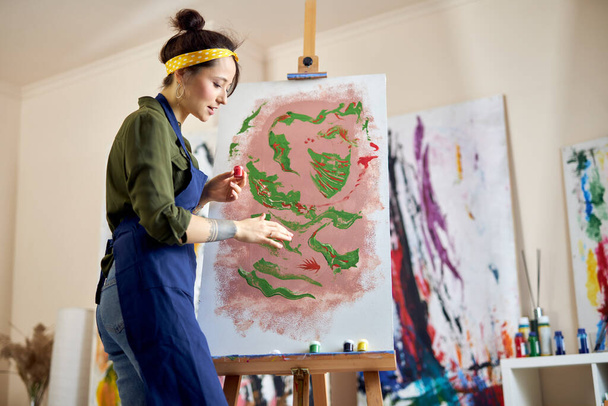 Вид збоку молодої художниці в фартусі, що малює картину на мольберті, наносить фарбу на полотно з пальцями, що стоять у вітальні в домашній студії
 - Фото, зображення
