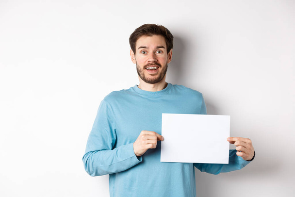 Romantic man looking with hopeful face at camera, showing sign card, blank piece of paper for banner or logo, standing over white background - Zdjęcie, obraz