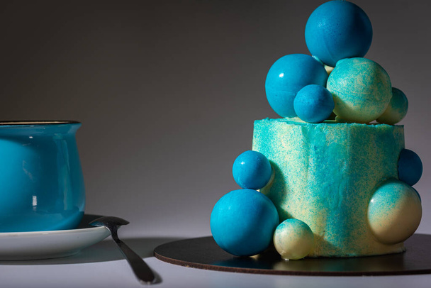 Gâteau d'anniversaire bleu avec des boules de chocolat blanc et bleu avec une tasse de café bleu, sur un fond gris. Ombre et lumière, contraste - Photo, image