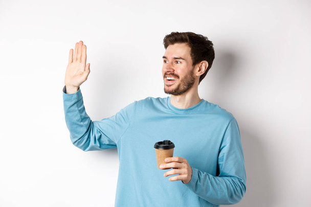 Friendly young man drinking coffee from takeaway cup and saying hello, waving hand left and smiling, standing over white background - Photo, Image
