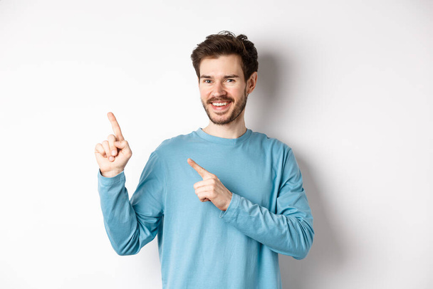 Cheerful bearded man showing advertisement on copy space, pointing fingers at upper left corner logo and smiling, standing over white background - Photo, Image