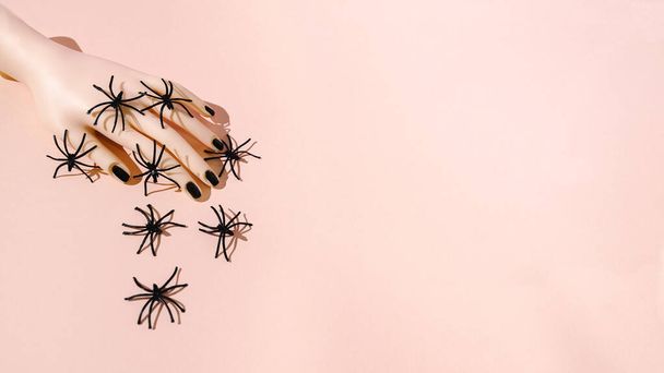 Eine Hand mit schwarzen Nägeln, die mit Spinnen bedeckt sind. Pastellrosa Hintergrund. Horror-Hintergrund zu Halloween oder kreatives Design. Kopierraum. - Foto, Bild