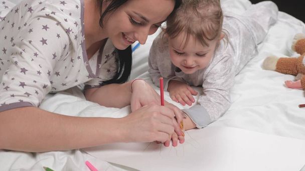 Anya, dadus, megtanítja a lányt rajzolni, a tenyér ceruzával van körvonalazva. Boldog család játszik együtt otthon az ágyban. Anya segít a lányának megtanulni papírra rajzolni, színes ceruzákkal színezni. - Fotó, kép