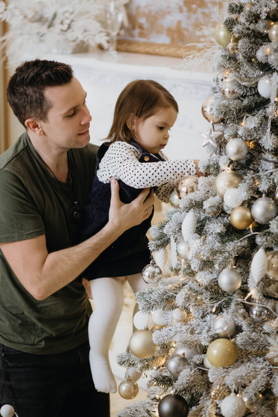 Père et jolie fille décorent le sapin de Noël. Papa tient soigneusement sa fille dans ses bras et décore le sapin pour les vacances de Noël. Nouvel An et vacances de Noël en famille - Photo, image