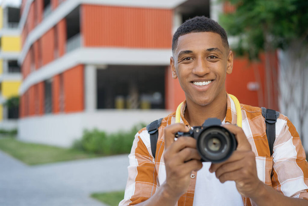 Portret van professionele Afro-Amerikaanse fotograaf die foto 's maakt op straat terwijl hij naar de camera kijkt. Toerist met digitale camera buiten, kopieerruimte. Reisconcept - Foto, afbeelding