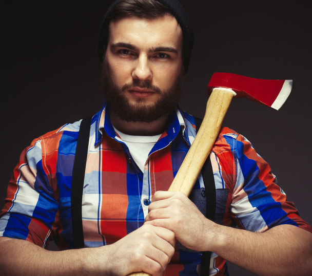 Bearded man wearing plaid shirt and balck hat with axe posing over black background in studio - Photo, image