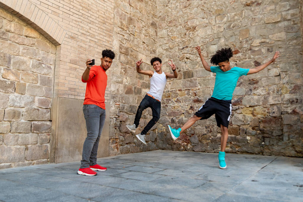 3人のアフロラテン系の男性の友人が路上で踊り、それを記録する - 写真・画像