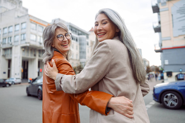 Happy senior Asian woman and grey haired friend hug walking along city street - Photo, image