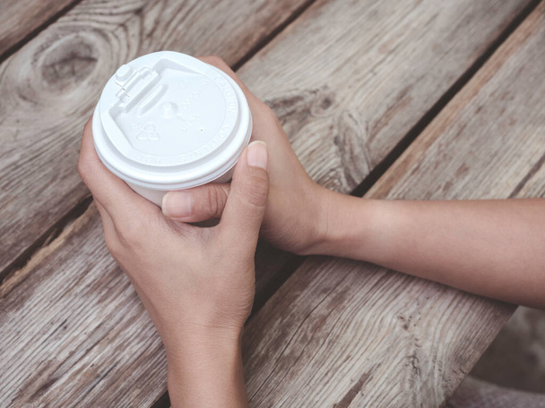 木製のテーブルの上に白いコーヒーペーパーカップを保持している女性の手を閉じます。使い捨てカップからコーヒーを飲む若い女性. - 写真・画像
