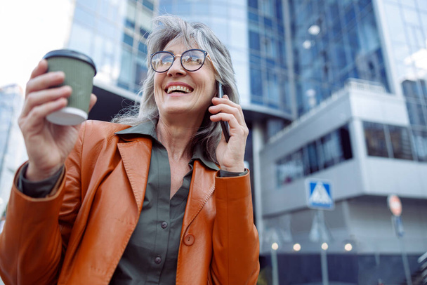 Happy female pensioner with cup of drink n talks on mobile phone on modern city street - Fotó, kép