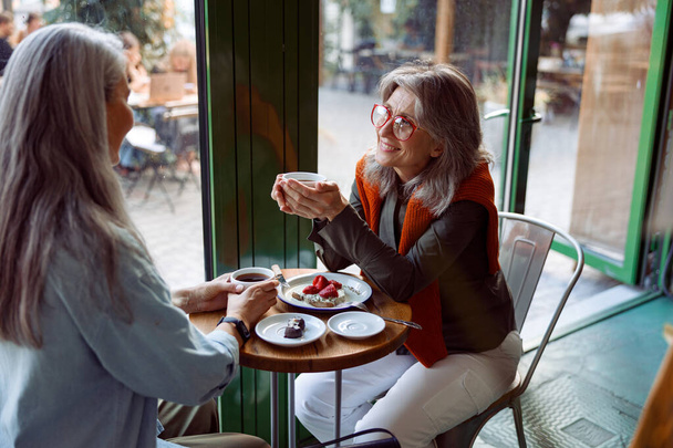 Позитивна зріла жінка і компаньйон з сірим волоссям насолоджуються напоями за столом у затишному кафе
 - Фото, зображення