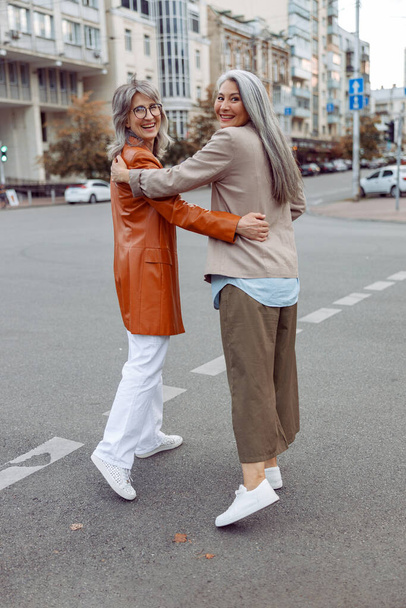 Lächelnde ältere asiatische Dame und grauhaarige Begleiterin umarmen sich beim Spazierengehen entlang der Stadtstraße - Foto, Bild