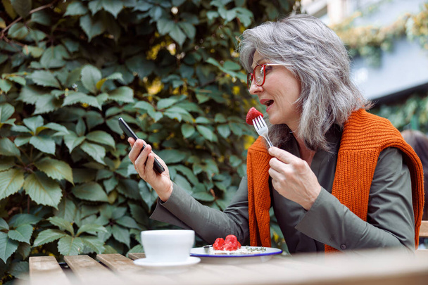 Mujer madura con gafas come postre y mira el teléfono inteligente en la mesa al aire libre - Foto, imagen