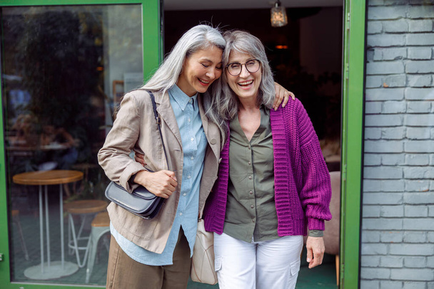 Pretty senior Asian lady hugs friend in purple jacket standing near cafe on street - Fotó, kép