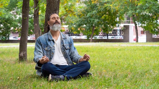 Foto di un uomo barbuto di mezza età che si prende una pausa dal trambusto della città. Sta meditando nel parco sotto un albero. Concetto di disintossicazione digitale. Formato foto 16x9. - Foto, immagini
