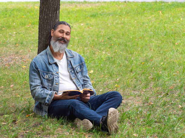Mann mittleren Alters mit einem Buch im Park. Er sitzt auf dem Rasen im Park und blickt in die Kamera. Digitales Entgiftungskonzept. - Foto, Bild