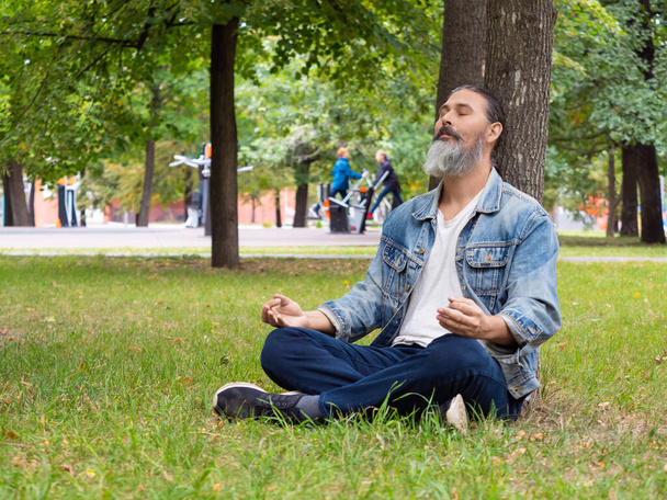 Egy szakállas középkorú férfi, aki szünetelteti a város nyüzsgését. Meditál a parkban egy fa alatt. Digitális méregtelenítés. - Fotó, kép