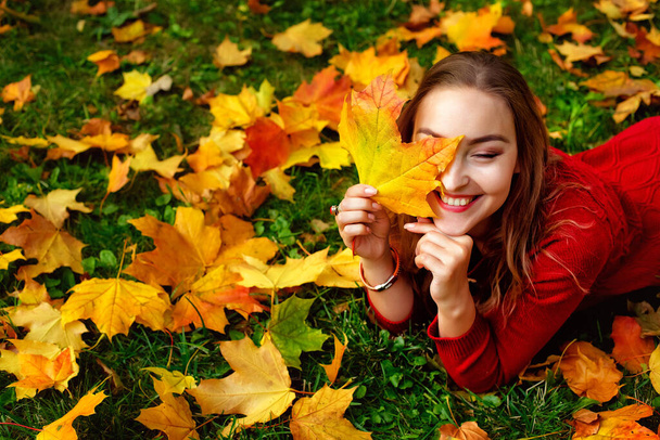 美しい公園で、秋の黄色と赤の葉で覆われた地面に横たわって、赤いニットのドレスで陽気な若い女性。彼女は目を閉じて、彼女の顔にモミジの葉を入れた。. - 写真・画像