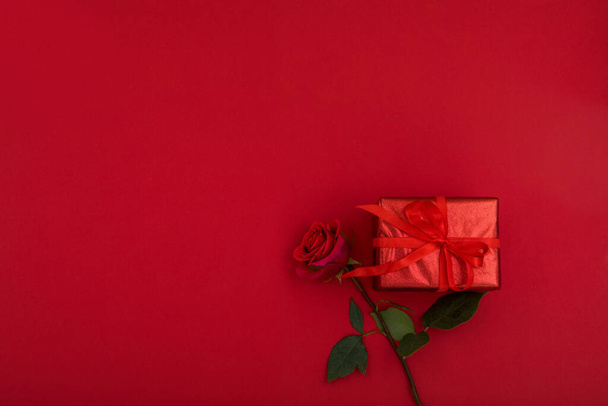 Вид зверху невелика подарункова коробка, загорнута червоним папером зі стрічкою, розміщеною поблизу квітучої троянди на яскраво-червоному тлі з порожнім простором для романтичного дизайну листівки на день Святого Валентина
 - Фото, зображення