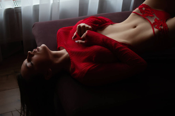 High angle side view of sensual young female in red knitted sweater and sexy lace red panties lying on bed - Photo, Image