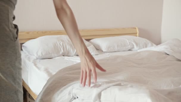 Nainen harmaissa housuissa peittää suuren sängyn valkoisella pehmeällä peitolla pitäen järjestystä valoisassa huoneessa lähikuvassa. Kotitaloudet ja arkiaskareet - Materiaali, video