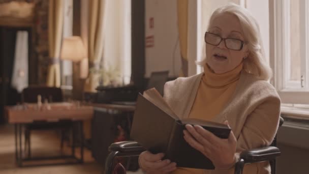 Plan moyen de vieille dame intelligente en fauteuil roulant lisant à haute voix puis discutant du livre avec une jeune infirmière soignante à la maison pour les personnes âgées - Séquence, vidéo