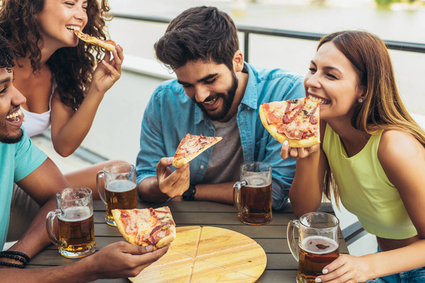 Přátelé si užívají pizzu. Skupina mladých veselých lidí jíst pizzu a pít pivo, zatímco sedí u fazolových pytlů na střeše budovy - Fotografie, Obrázek