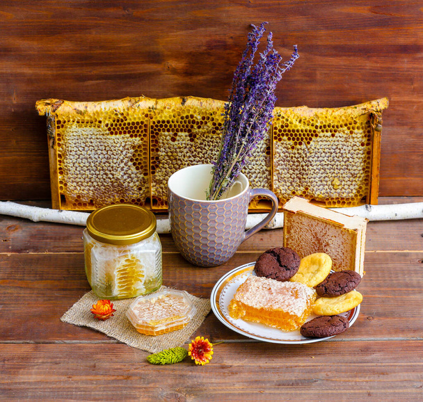 Rustic still life with beekeeping products: honey combs in a glass jar, wild honey in a honeycomb frame on a birch branch, a cookies and cut combs on a plate, on a wooden background - Foto, Imagen
