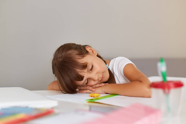 Indoor shot of little schoolgirl sleeping while sitting at the table, being tired while doing doing her homework, child with dark hair wearing white t shirt. - Zdjęcie, obraz