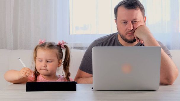 padre divertido de un hombre de negocios está trabajando en una computadora portátil en casa junto a una niña, 3 años de edad, jugando en la tableta. Familia moderna. Concepto independiente. Adicción a los aparatos - Foto, Imagen