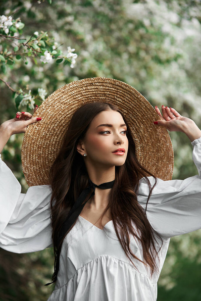 Brown-haired woman in a large straw hat and a white dress poses against the background of blooming white trees. Romantic look, natural beauty, clean facial skin - Foto, Imagen