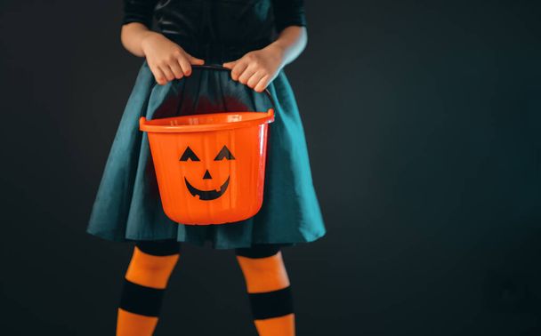Joyeux Halloween ! Petite fille mignonne en costume de sorcière avec panier pour bonbons sur fond de mur noir. - Photo, image