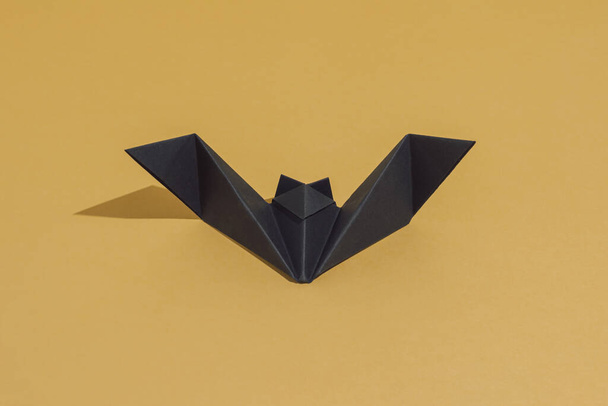 Nietoperz Origami wykonany z czarnego papieru, izolowany na tle złota lub ochry. Zrób to sam pomysł dekoracji z miejsca na ksero. Słońce i cienie. Minimalne luksusowe zaproszenie na imprezę Halloween. - Zdjęcie, obraz
