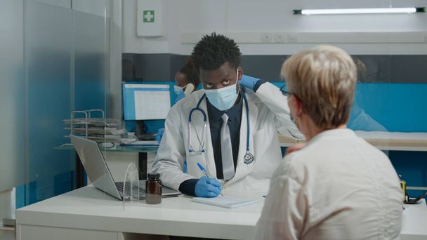 Médico con abrigo blanco sentado en el escritorio mientras habla con la mujer mayor - Foto, imagen