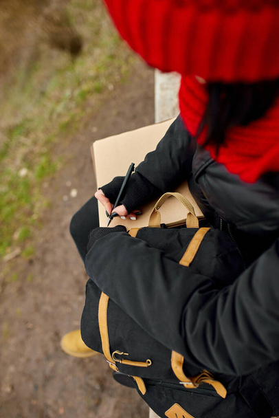 Mujer de viaje con sombrero rojo en chaqueta negra escribir en el cuaderno, tomar notas, dibujar algunos bocetos contra la vista del río, los viajes locales, la salud mental. Otoño, otoño - Foto, Imagen