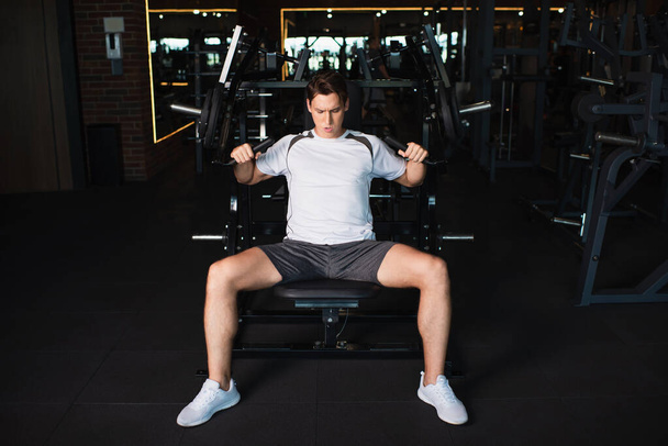 vista completa del hombre atlético haciendo ejercicio en la máquina de ejercicios de extensión de brazo en el gimnasio - Foto, imagen