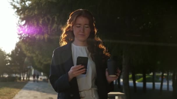 Позитивная молодая женщина держит кубок и мобильный телефон в городском парке - Кадры, видео