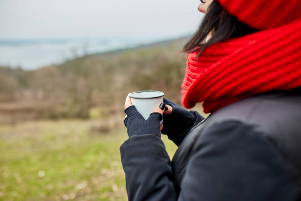 Kırmızı şapkalı ve eşarplı kadın, metal bardaktan çay içiyor, seyahat konsepti, sonbahar manzarası. - Fotoğraf, Görsel