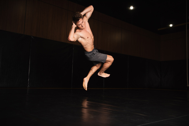 shirtless and barefoot sportsman in shorts jumping with hands behind head in gym - Photo, Image