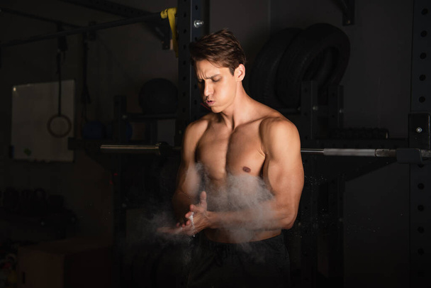 sin camisa deportista muscular hinchando mejillas mientras se aplica talco en polvo en las manos - Foto, Imagen