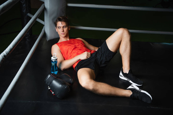 tired sportsman resting in corner of ring near boxing gloves and sports bottle - Photo, Image