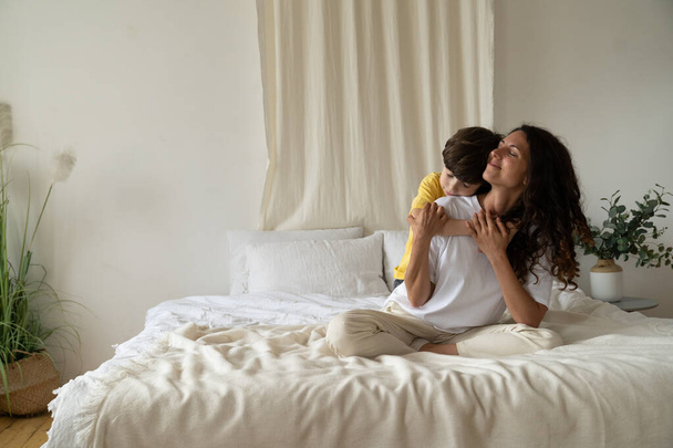 Il ragazzo abbraccia la mamma sorridente al mattino seduta sul letto con il pigiama a casa il fine settimana. Amore familiare - Foto, immagini